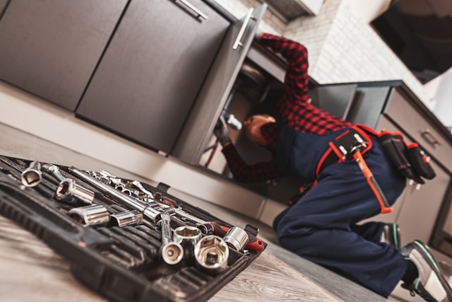 Male Neighborhood Plumber wearing a red checkered shirt and blue overalls, under the kitchen sink, with a close up of his tool box.