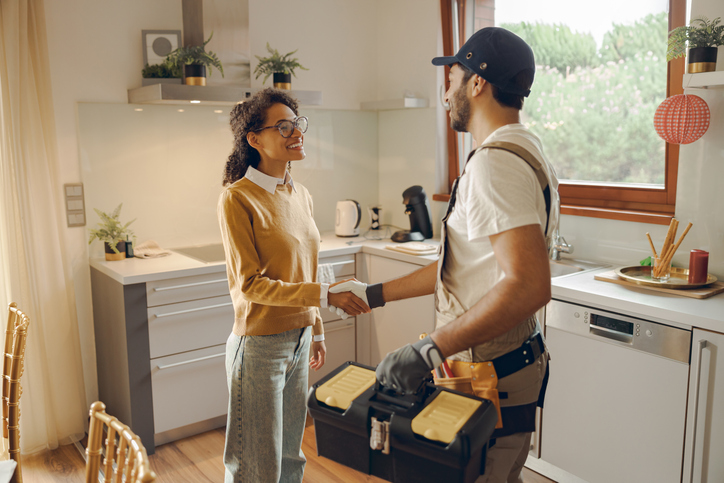A Delta Neighborhood Plumber is shaking hands with his new client. She is a young person of colour, wearing glasses, a mustard coloured shirt and mom jeans. They are standing in the kitchen of the clients house.