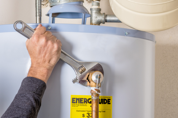 A plumber wearing a long sleeve dark grey shirt is using a wrench to tighten a pipe on a hot water tank. Hot Water Tank Replacement in Delta