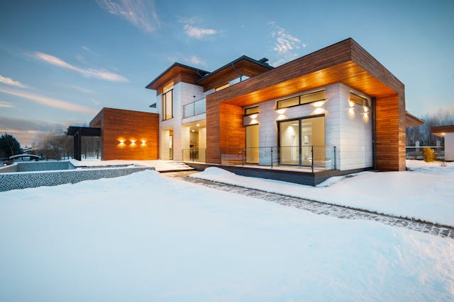 A beautiful custom home with white and brown details, lots of windows and lights. The ground is covered in snow. Protecting Your Pipes During the Winter