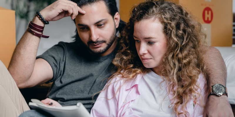 a white man with long dark hair pulled back, a dark bread is with his wife, she is wearing a pink and white shirt and has long curly brown hair. they are reading an Emergency Plumbing Checklist and have boxes around them.