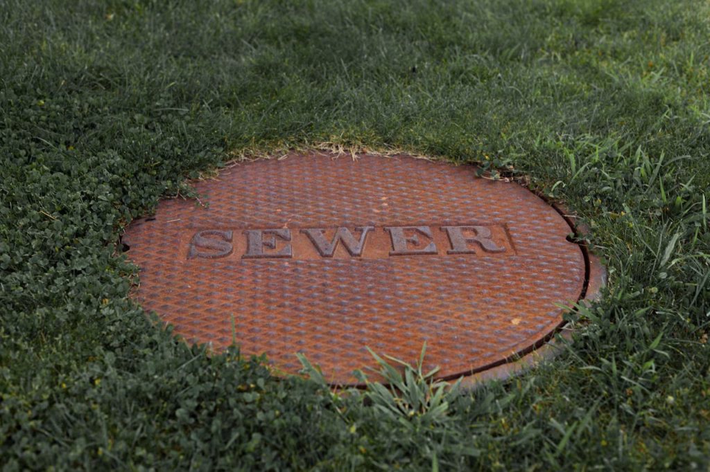 A brown sewer lid in a yard of grass. It says Sewer across the top. Camera Inspection entry. 